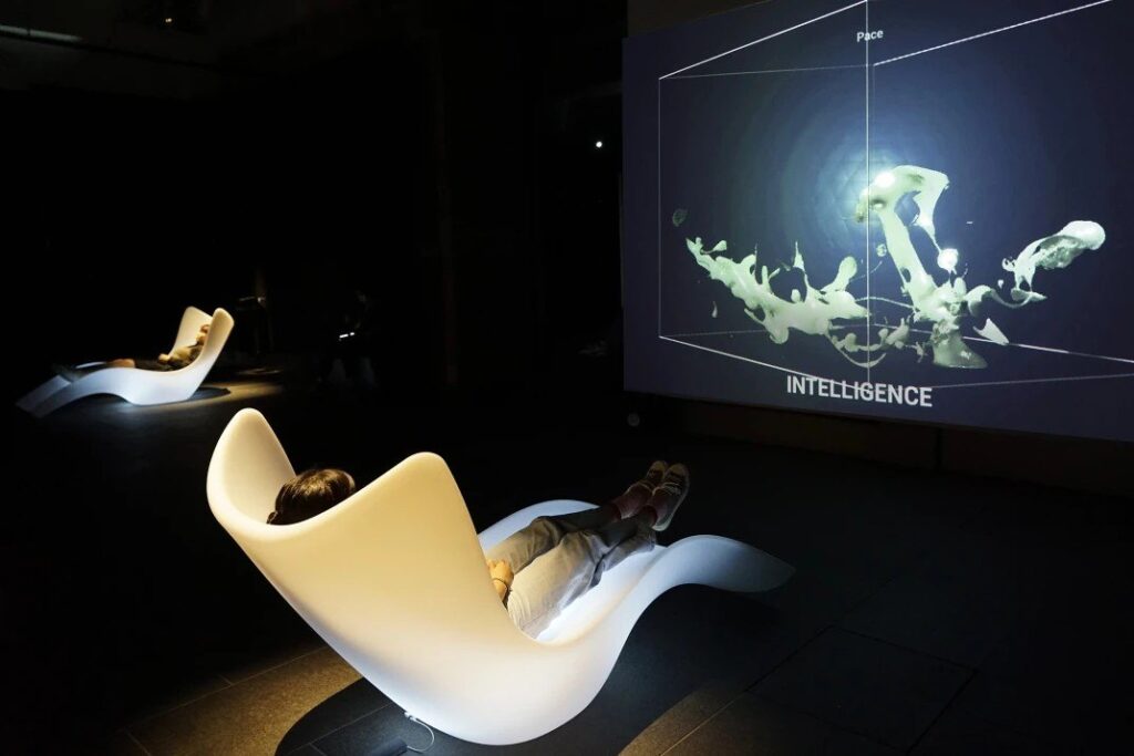 Visitor reclining in a white lounge chair at Maurice Benayoun's Neuro-Design Station while a 3D form labeled Intelligence is projected on the wall
